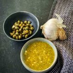 Caper Vinaigrette in a small bowl next to a bowl of capers and whole garlic