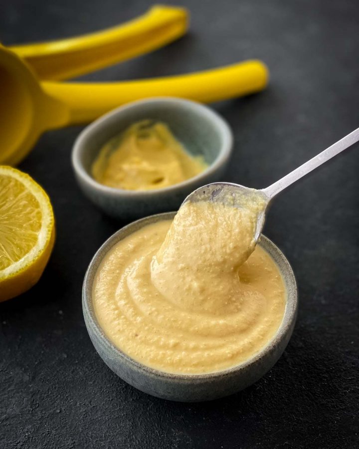 A teaspoon in a small bowl of Maple Hummus Dressing with mustard and a lemon squeezer in the background