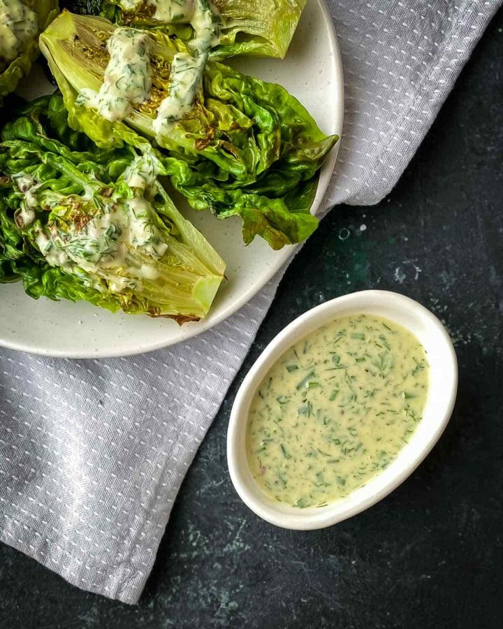 Tahini Ranch Dressing in a small white bowl sitting next to grilled cos lettuce with the plate resting on a grey napkin.