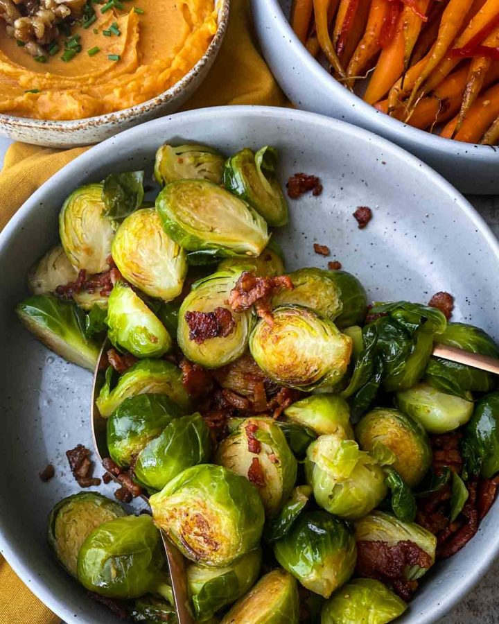 Brussels Sprouts With Bacon in a grey bowl with serving cutlery served with whipped sweet potatoes and roasted carrots on the side