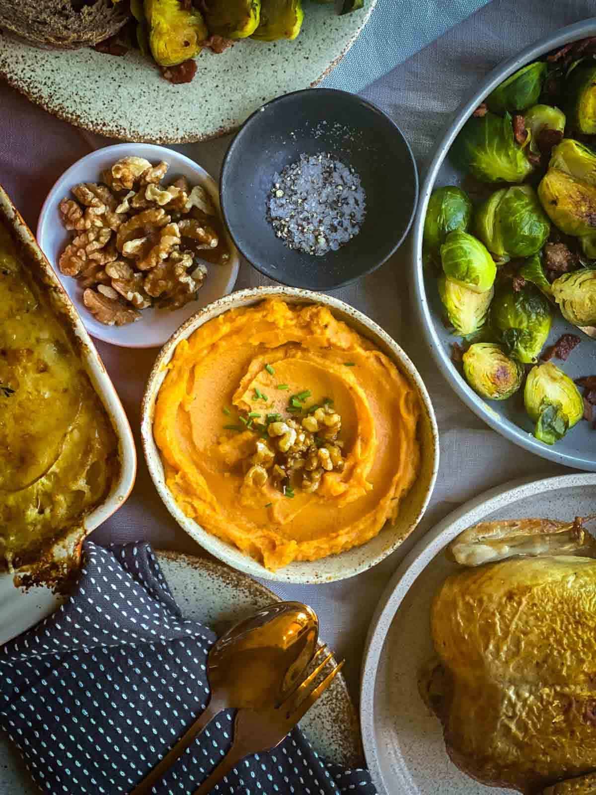 A spoon in a bow of whipped sweet potato next to a bowl of walnuts, Brussel sprouts and roast chicken