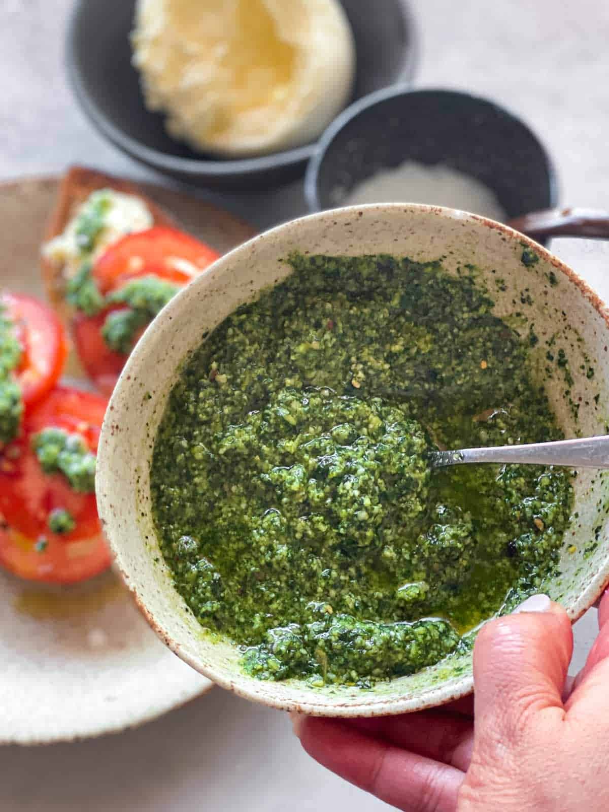 Hand holding a bowl of basil mint pesto hovering over sliced tomatoes and burrata