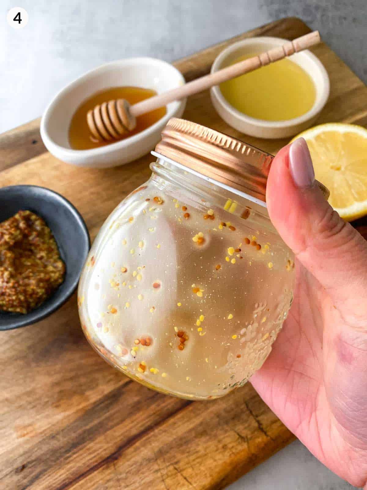 Shaking a jar of Honey Seeded Mustard Dressing, hovering over the ingredients sitting on a wooden board