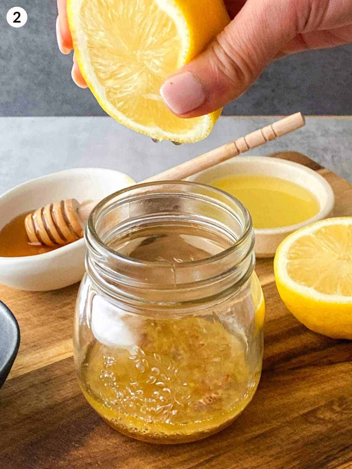 Squeezing half a lemon into a jar of olive oil and seeded mustard with honey and lemon in the background