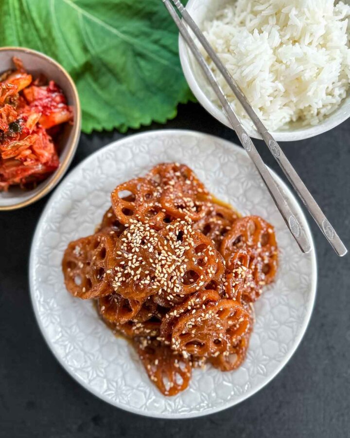 Korean Braised Lotus Roots in a white plate with chopsticks served with white rice, kimchi and perilla leaves in the background